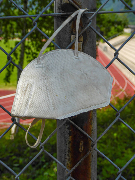 stara, brudna i porzucona maska FFP2 wisi na płocie w pobliżu publicznego boiska sportowego. Symbol odpadów i zanieczyszczenia - Zdjęcie, obraz