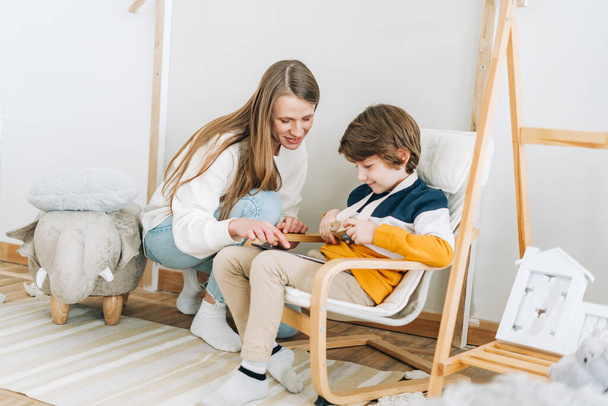 Happy caucasian mother sitting with son in childrens room, looking at tablet computer device. Smiling family plays video games with kid boy together. Mom and chilld wathcing on touch screen - Foto, Imagem