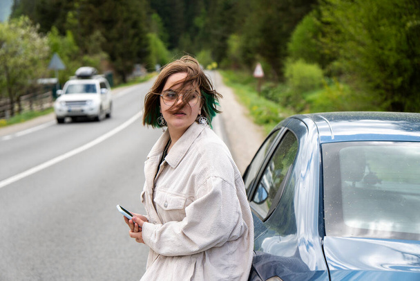 Портрет красивой молодой женщины со сломанной машиной на дороге, держащей телефон - Фото, изображение