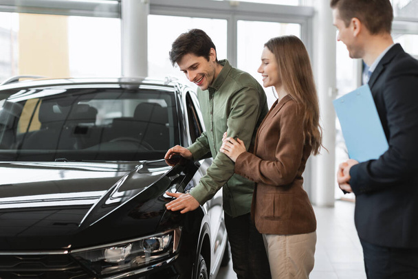 Millennial Caucasian couple choosing new car, communicating with auto salesman at vehicle dealership - Fotoğraf, Görsel