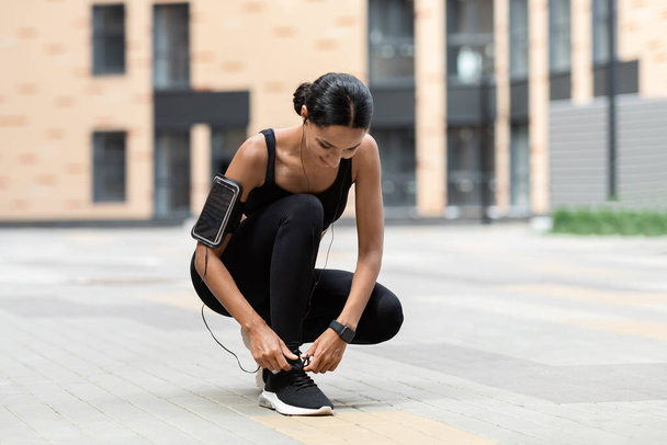 Lachende jonge islamitische dame in sportkleding met telefoon op de schouder, fitness tracker strikken schoenveters klaar om te rennen - Foto, afbeelding