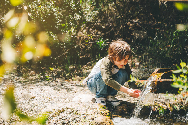 School boy kid drinking water from the mountain creek. Tourist child wearing casual clothes making a sip of mountain river water from the palms of his hands when hiking - Foto, Imagen