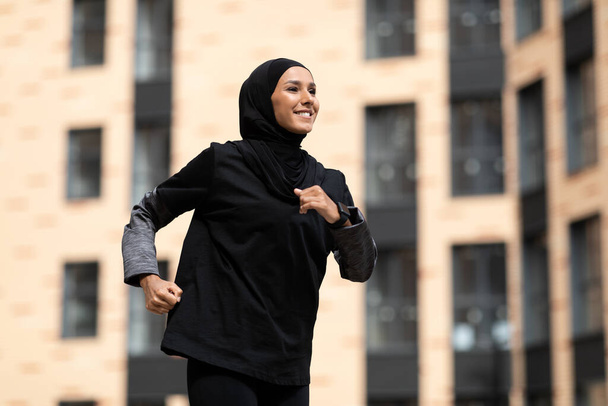 Ευτυχής χαμογελαστός νεαρός Άραβας γυναίκα σε μαντίλα με tracker φυσικής κατάστασης τρέχει σε φόντο κτιρίου - Φωτογραφία, εικόνα