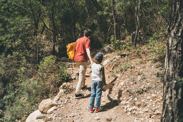 Rear view of tourists school boy and his dad walking a stone footpath in spring forest. Child kid and father wearing casual clothes hiking in summer greenwood leaf forest - Photo, Image
