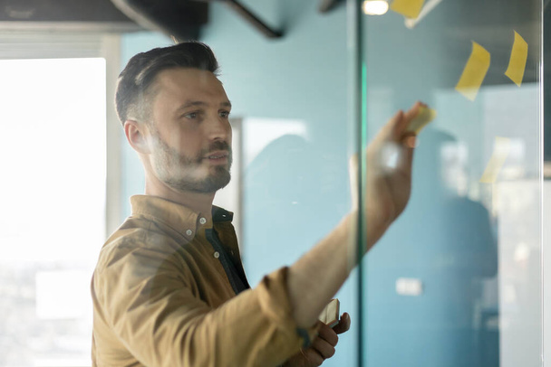 Männlicher Unternehmer klebt Klebezettel an Glaswand im Büro - Foto, Bild