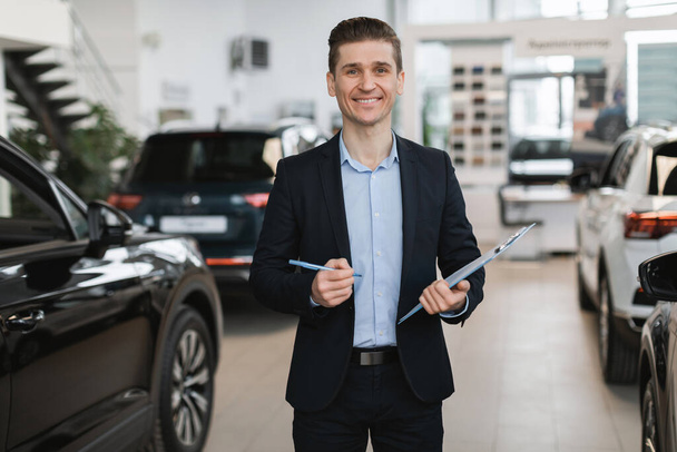 Portrét pozitivního mladého agenta automobilového pojištění pózujícího v prodejně automobilů - Fotografie, Obrázek