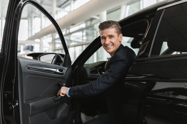 Успешный продавец автомобилей тысячелетия сидит внутри автосалона, смотрит в камеру через открытую дверь в современном салоне - Фото, изображение