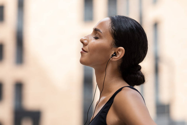 Glad young arab female with closed eyes in headphones enjoy music and relaxing on building background outdoor, profile - 写真・画像