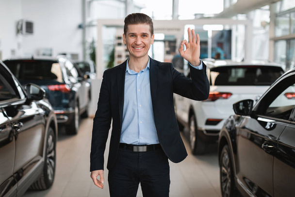Uśmiechnięty sprzedawca samochodów wykazujący się dobrym gestem, stojący wśród nowych samochodów, rekomendujący nowoczesny salon samochodowy - Zdjęcie, obraz