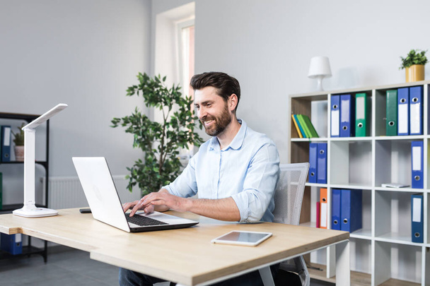 Portrait of successful businessman, man smiling and working on laptop in modern bright office - Φωτογραφία, εικόνα