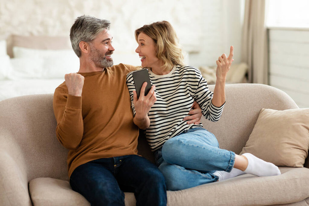 Fröhliches Paar benutzt Handy und gestikuliert zu Hause - Foto, Bild