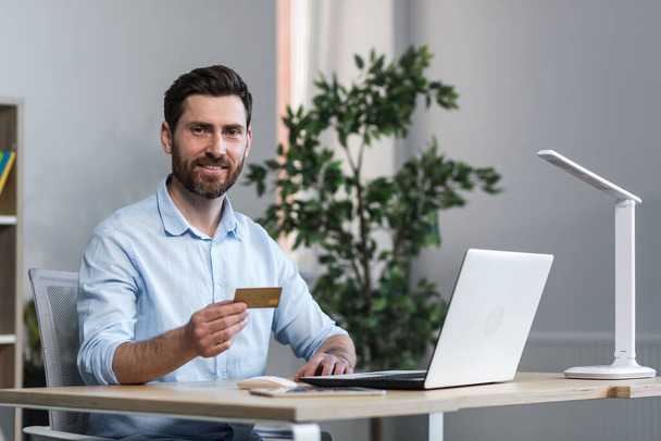 Happy businessman makes online purchases working in the office, husband holds credit bank card and uses laptop - Foto, Imagem