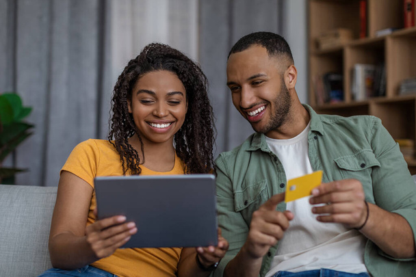 Щасливі чорні подружжя купують в Інтернеті за допомогою планшета і кредитної картки, платять за замовлення, сидячи на дивані вдома
 - Фото, зображення