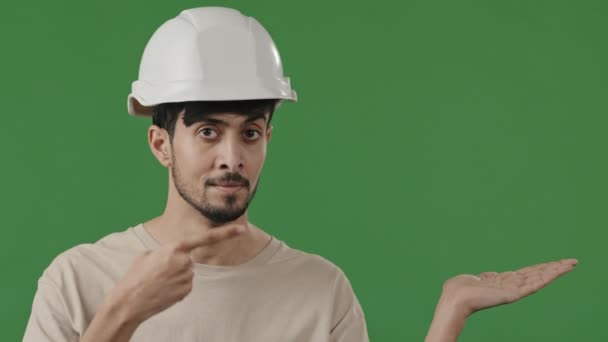 Portret pewny siebie arabski człowiek mechanik mężczyzna inżynier nosić specjalny kask bezpieczeństwa patrząc na aparat uśmiechnięty hiszpański inżynier brygadzista inspektor stwarzając punkt wskazujący palec na prawej ręce w zielonym studio - Materiał filmowy, wideo
