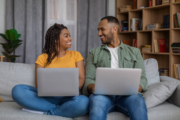 Joven pareja negra usando computadoras portátiles, mirándose y sonriéndose, sentándose en el sofá y navegando por Internet - Foto, Imagen