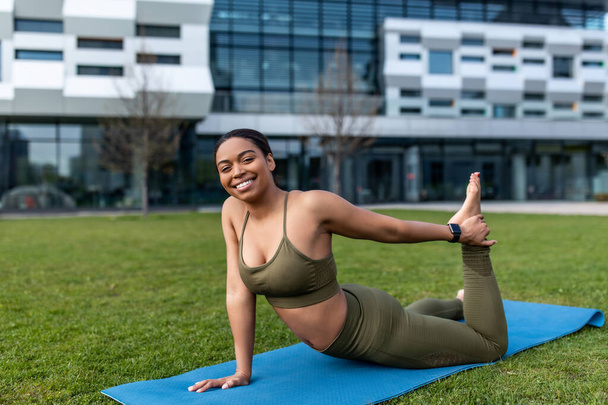 Athletic young black woman in sportswear doing fitness exercises on yoga mat, stretching her leg at city park - Photo, Image