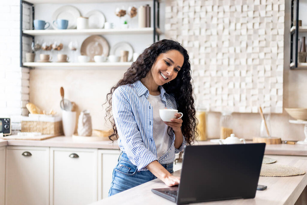 Onnellinen latino nainen käyttää kannettavaa tietokonetta ja juo kuumaa aamukahvia, työskentelee etänä kotoa - Valokuva, kuva