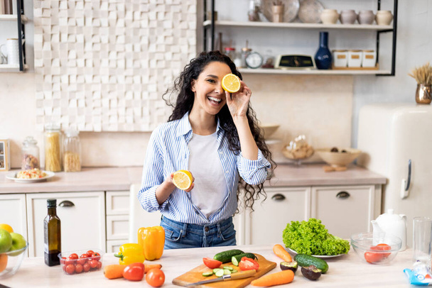 Jovem senhora latina feliz se divertindo e cobrindo os olhos com metades de limão, brincando na cozinha enquanto cozinha o jantar - Foto, Imagem