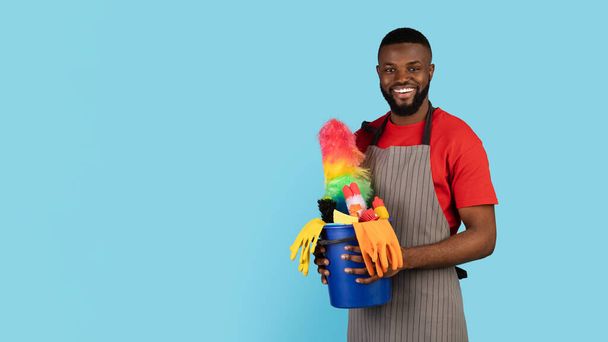Siivouspalvelut. Komea musta siivooja mies poseeraa pesuaineiden korilla - Valokuva, kuva