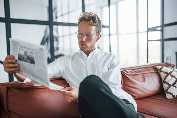 Reads newspaper. Caucasian young guy in elegant white shirt indoors at home. - 写真・画像