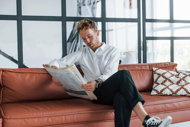 Reads newspaper. Caucasian young guy in elegant white shirt indoors at home. - Foto, Imagen