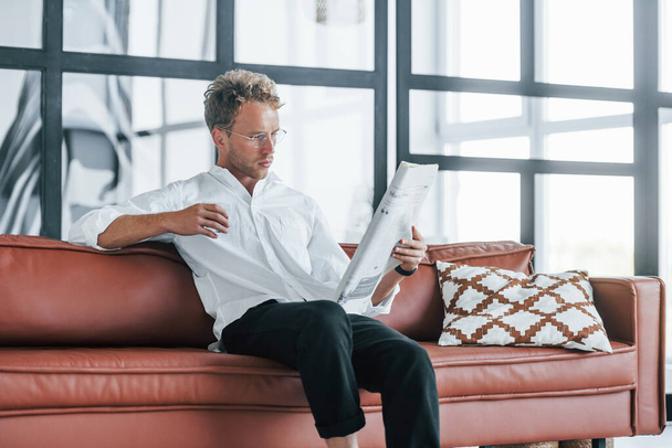 Reads newspaper. Caucasian young guy in elegant white shirt indoors at home. - Photo, image