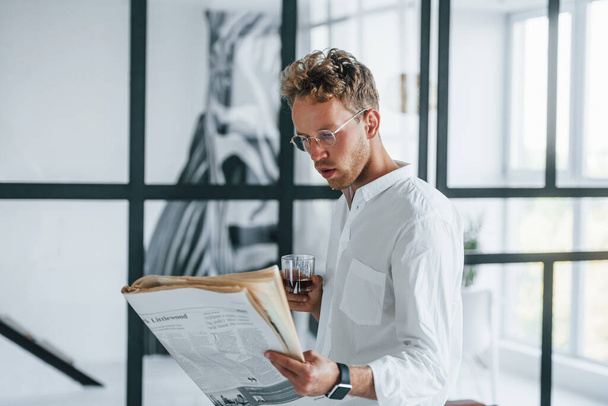 Reads newspaper. Caucasian young guy in elegant white shirt indoors at home. - Foto, Imagem