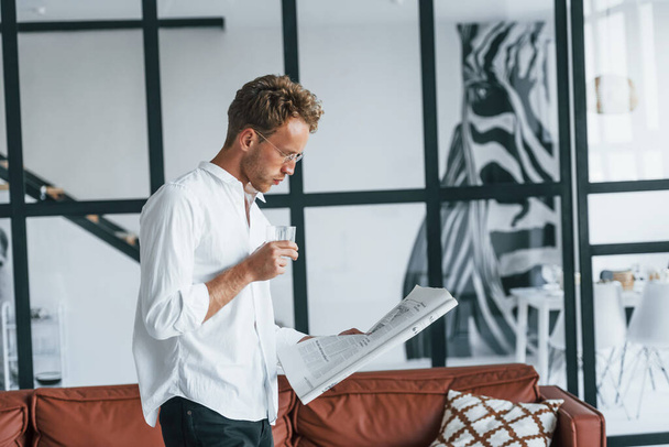 Reads newspaper. Caucasian young guy in elegant white shirt indoors at home. - Foto, afbeelding