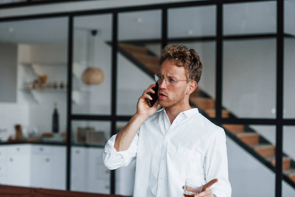 Talks by phone. Caucasian young guy in elegant white shirt indoors at home. - Photo, Image
