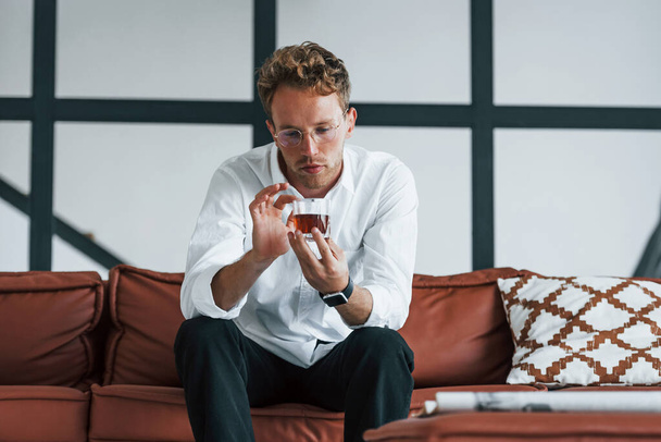 With drink in hands. Caucasian young guy in elegant white shirt indoors at home. - Fotoğraf, Görsel