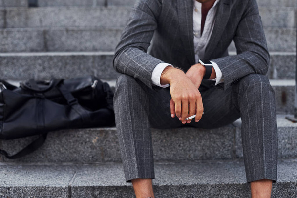 Sits and smokes cigarette. Young successful businessman in grey formal wear is outdoors in the city. - Fotoğraf, Görsel