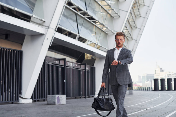 Checks time. Young successful businessman in grey formal wear is outdoors in the city. - Foto, afbeelding