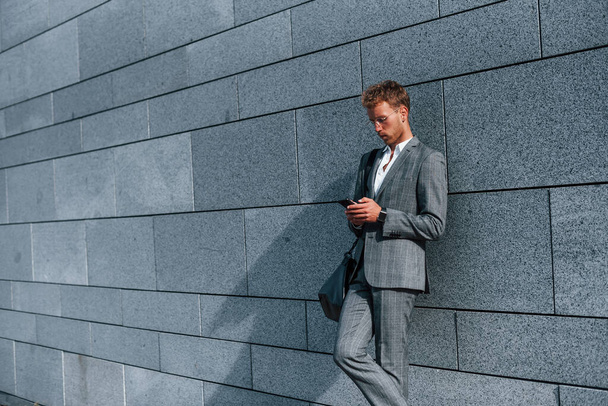 Leans on the wall. Young successful businessman in grey formal wear is outdoors in the city. - 写真・画像