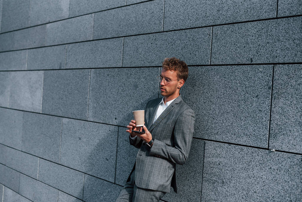 Leans on the wall. Young successful businessman in grey formal wear is outdoors in the city. - Foto, afbeelding
