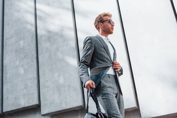 In sunglasses. Young successful businessman in grey formal wear is outdoors in the city. - Foto, Bild