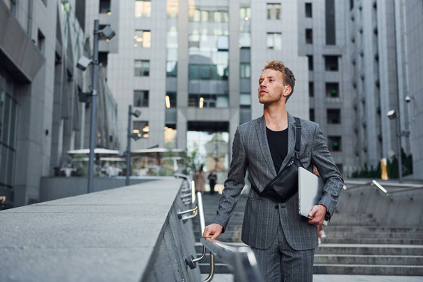 Laptop in hands. Young elegant man in good clothes is outdoors in the city at daytime. - 写真・画像