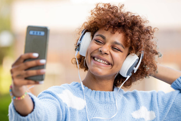 menina americana africana com fones de ouvido e fones de ouvido fazendo foto ou vídeo com telefone celular - Foto, Imagem
