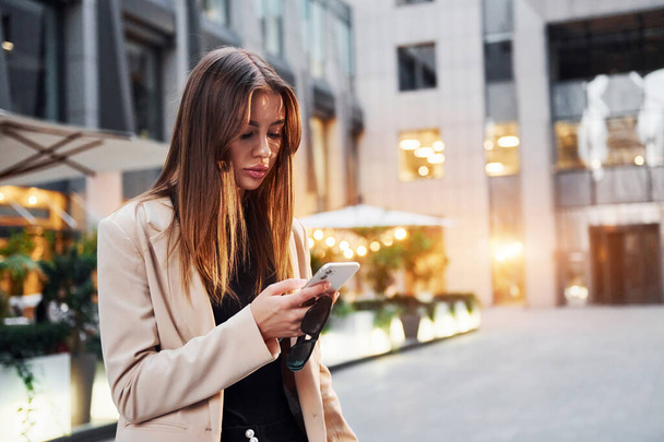Uses phone. Beautiful young european woman is outdoors in the city at daytime. - 写真・画像