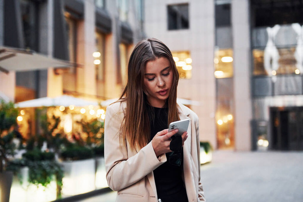 Uses phone. Beautiful young european woman is outdoors in the city at daytime. - Foto, Bild