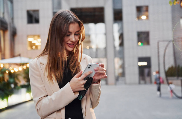 Uses phone. Beautiful young european woman is outdoors in the city at daytime. - Фото, зображення