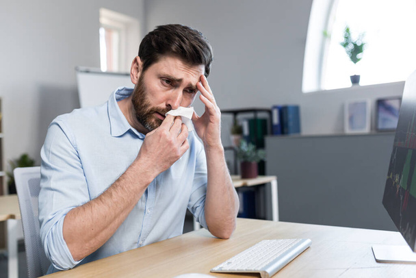 Sick and tired young man at work. Office worker, manager, freelancer sitting at a desk in the office does not feel well, wipes his nose, has a runny nose, coughs, headaches, holds his head. - 写真・画像