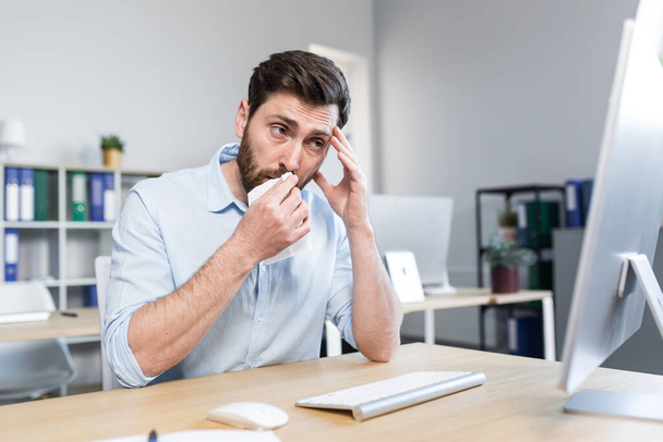 Sick and tired young man at work. Office worker, manager, freelancer sitting at a desk in the office does not feel well, wipes his nose, has a runny nose, coughs, headaches, holds his head. - Photo, image