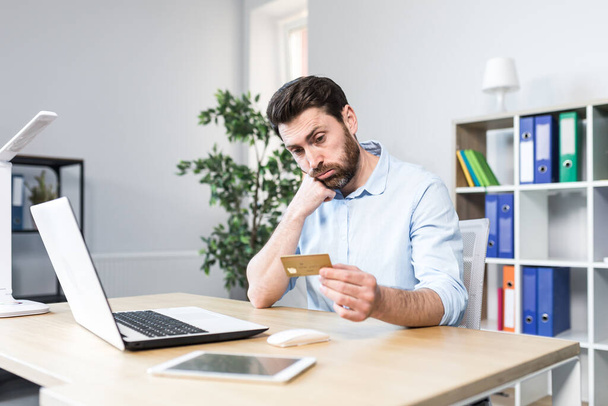 Unsuccessful online purchase. Unsuccessful deal. Upset young man, office worker holding a credit card, holding his head in shock. Sitting at a desk, at a laptop - Photo, Image
