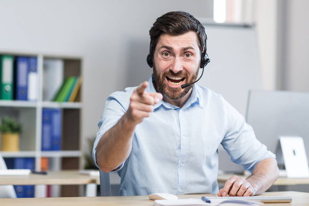 A young man, a consultant, an office worker, a manager talking on a video call in headphones, waving his arms, explaining something. Sitting at a computer desk in the office, in a white shirt - 写真・画像