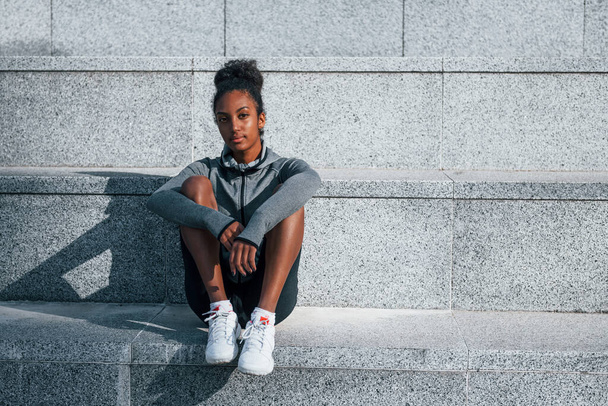 Sits on stairs. Young african american woman in sportive clothes have workout outdoors at daytime. - Photo, image