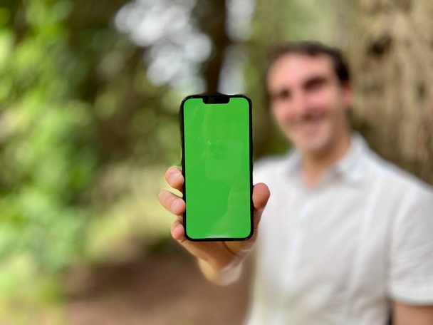 フォーカスのみの携帯電話フォーカスは非常にハンサムな男フランスイタリア白のシャツで森の中に立って緑のタップで電話を保持していないと笑みを浮かべて画面上の指を示すために使用することができます - 写真・画像