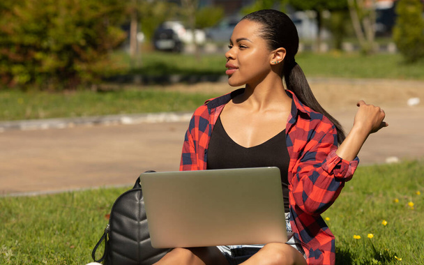 Joyful casual african-american student girl with laptop outdoors. Smiling woman sitting on grass with computer, surfing the net or preparing for exams.   - 写真・画像