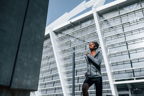 Big building behind. Young african american woman in sportive clothes have workout outdoors at daytime. - Photo, Image