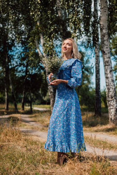Egy fiatal, gyönyörű nő kék ruhában vad virágokat tart a kezében. - Fotó, kép
