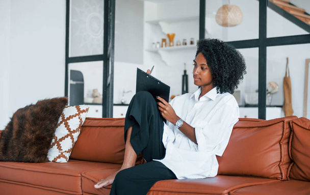 Holds notepad. Young african american woman with curly hair indoors at home. - Valokuva, kuva